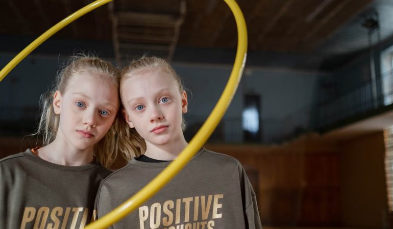 Twin girls in sportswear indoors with a hula hoop expressing positivity. Perfect for family and sports themes.